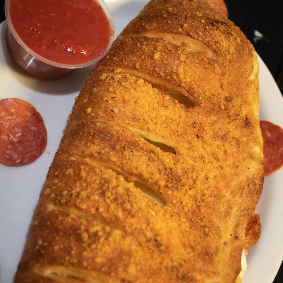 Golden-brown calzone with a side of red marinara sauce and pepperoni slices on a white plate.