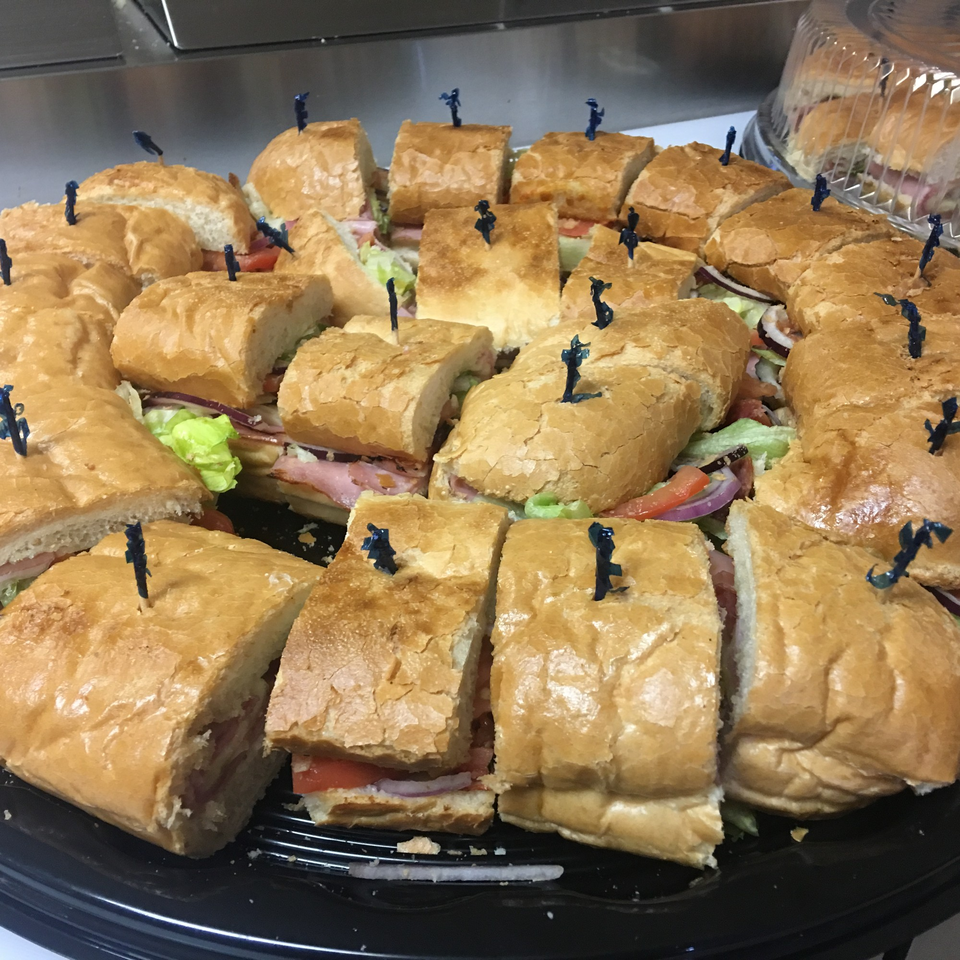 A catering platter of sliced submarine sandwiches with ham, lettuce, tomato, and onion.
